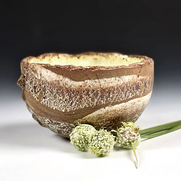 A handmade ceramic bowl with a rustic, textured stoneware finish in earthy brown and beige tones, styled with dried green flowers on a white surface against a dark gradient background.