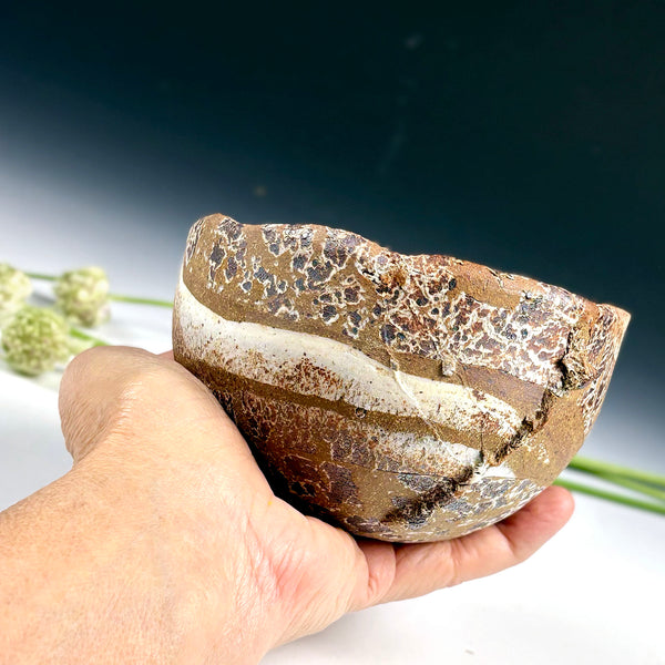 A hand holding a rustic handmade ceramic bowl with a textured, crackled brown and white glaze, set against a dark blue gradient background with blurred green stems.