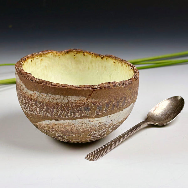 A handmade ceramic bowl with a rustic, uneven rim, featuring a textured brown and beige exterior and a pale cream interior, shown next to a silver spoon on a white surface with a dark gradient background.