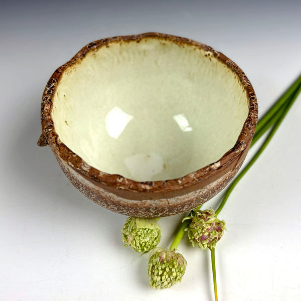 A handmade ceramic bowl with a rustic brown textured exterior and a glossy pale green glazed interior, styled with green flower buds on a white background.