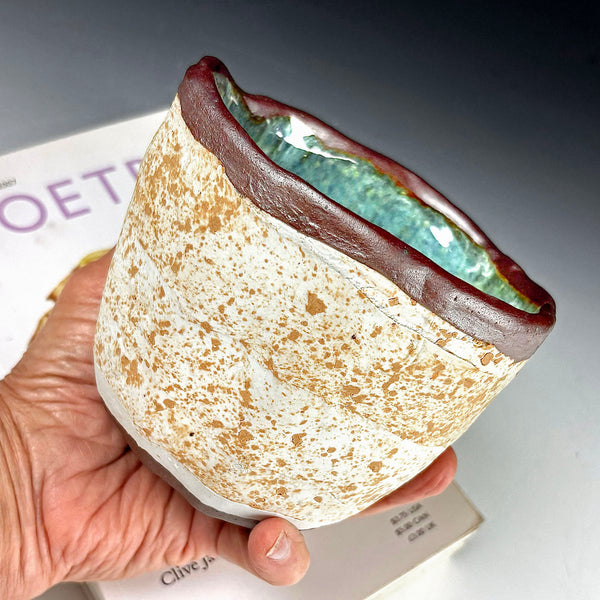 A handmade ceramic cup held in a hand, featuring a textured off-white body with gold speckles, a dark reddish-brown rim, and a glossy teal-green interior glaze.