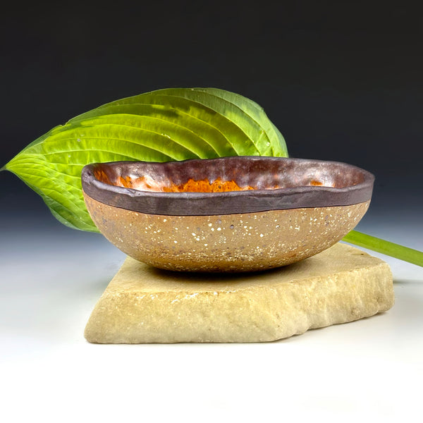 A handmade ceramic bowl with a speckled tan exterior and a dark brown glazed rim, displayed on a natural stone slab with a green leaf prop against a dark studio background.