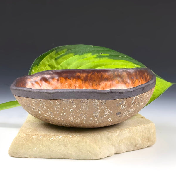 A handmade ceramic bowl with a textured speckled brown exterior and a glossy reddish-brown glazed interior, displayed on a light stone block with a green leaf behind it against a dark gradient background.