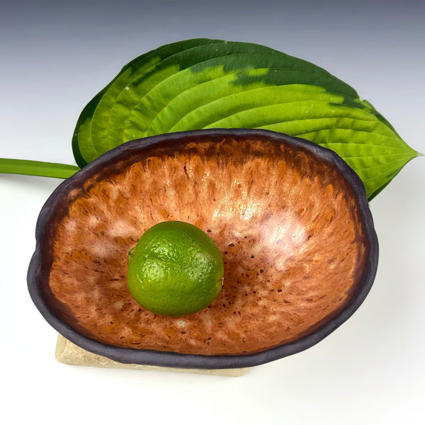 A handmade ceramic bowl with a textured brown interior and a dark glazed rim, shown on a white surface with a green lime in the center and a large green leaf behind it.