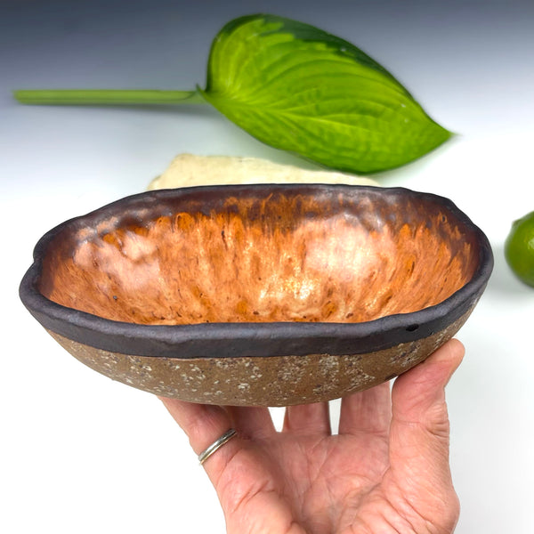 A handmade ceramic bowl with a textured, hammered metallic copper-orange interior and a dark brown rim, held by a hand against a white background with a green leaf in the background.