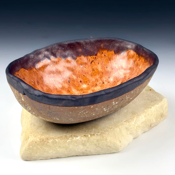 A handmade ceramic bowl with a textured orange and white interior and a dark glazed rim, displayed on a rough stone slab against a dark blue gradient background.