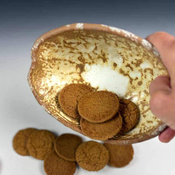 A hand holds a rustic, hand-painted ceramic bowl filled with round gingerbread cookies, with more cookies scattered on a white surface below.