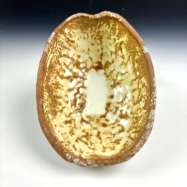 A handmade ceramic bowl with a rustic, organic shape, featuring a textured glaze with brown speckles and white patches on a light beige base, photographed against a neutral gradient background.
