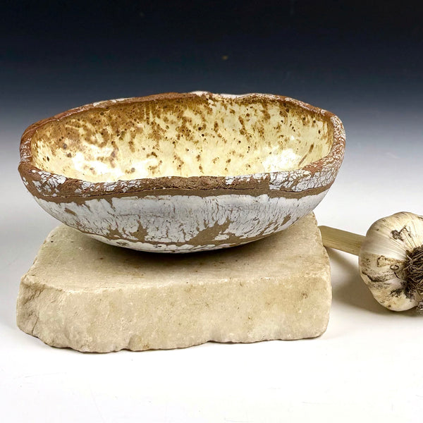 A rustic handmade ceramic bowl with a brown speckled interior and a white-glazed exterior, displayed on a stone block with a garlic bulb prop against a dark-to-light gradient background.