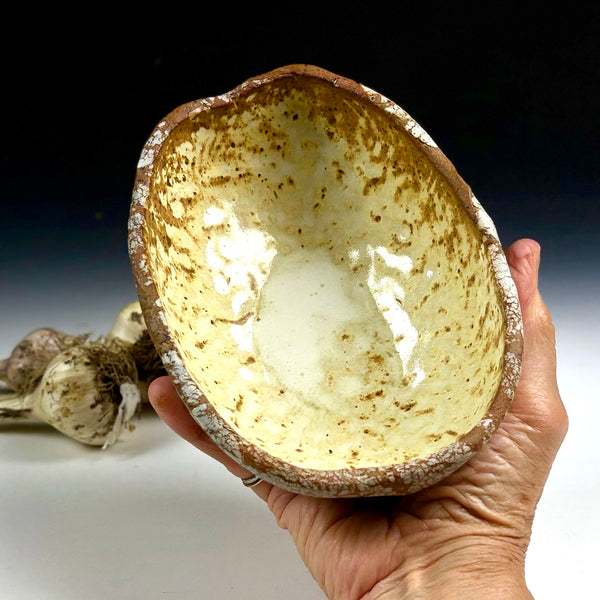 A handmade ceramic bowl with a rustic, organic shape, held in a person's hand. The bowl features a glossy cream-colored interior with brown speckled patterns and a rough, earthy brown exterior.