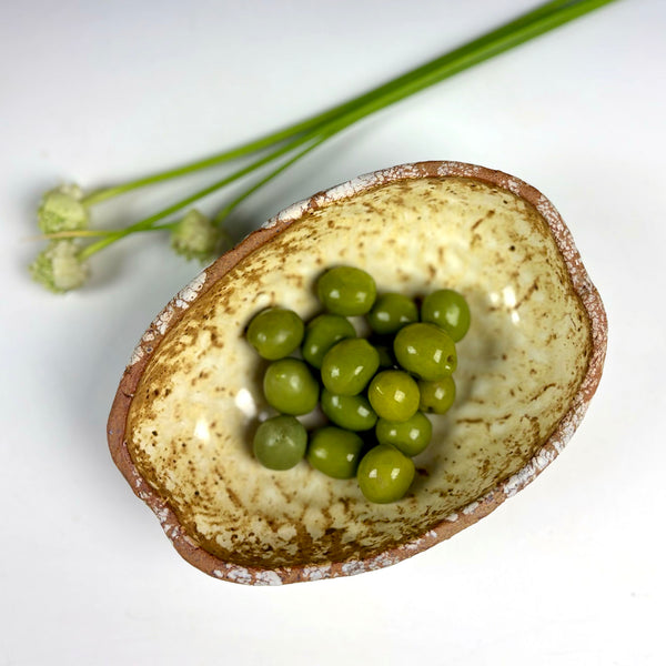 A small handmade ceramic bowl with a speckled beige and brown glaze, containing green peas, with dried flower stems in the background on a white surface.